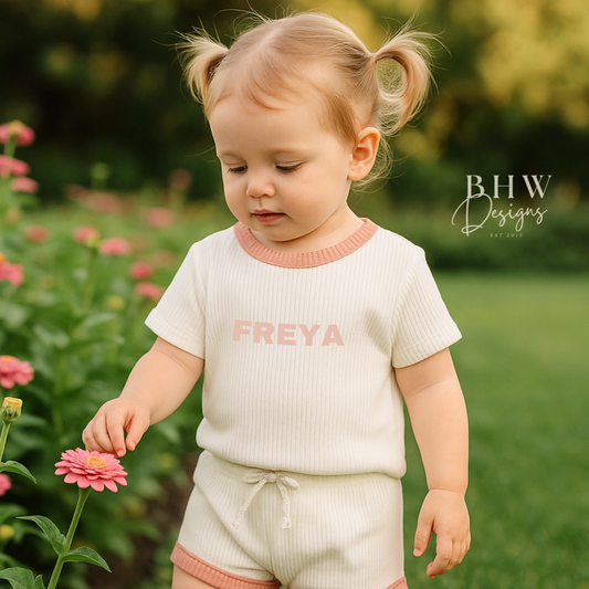 Child wearing a white ribbed outfit with baby pink trim and 'FREYA' printed on the front in baby pink. 