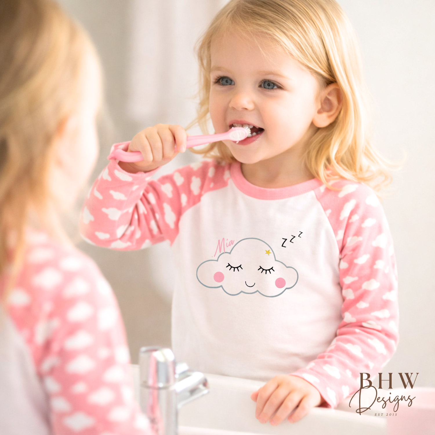 Child brushing teeth in front of a mirror wearing a pink and white shirt with a cloud design.