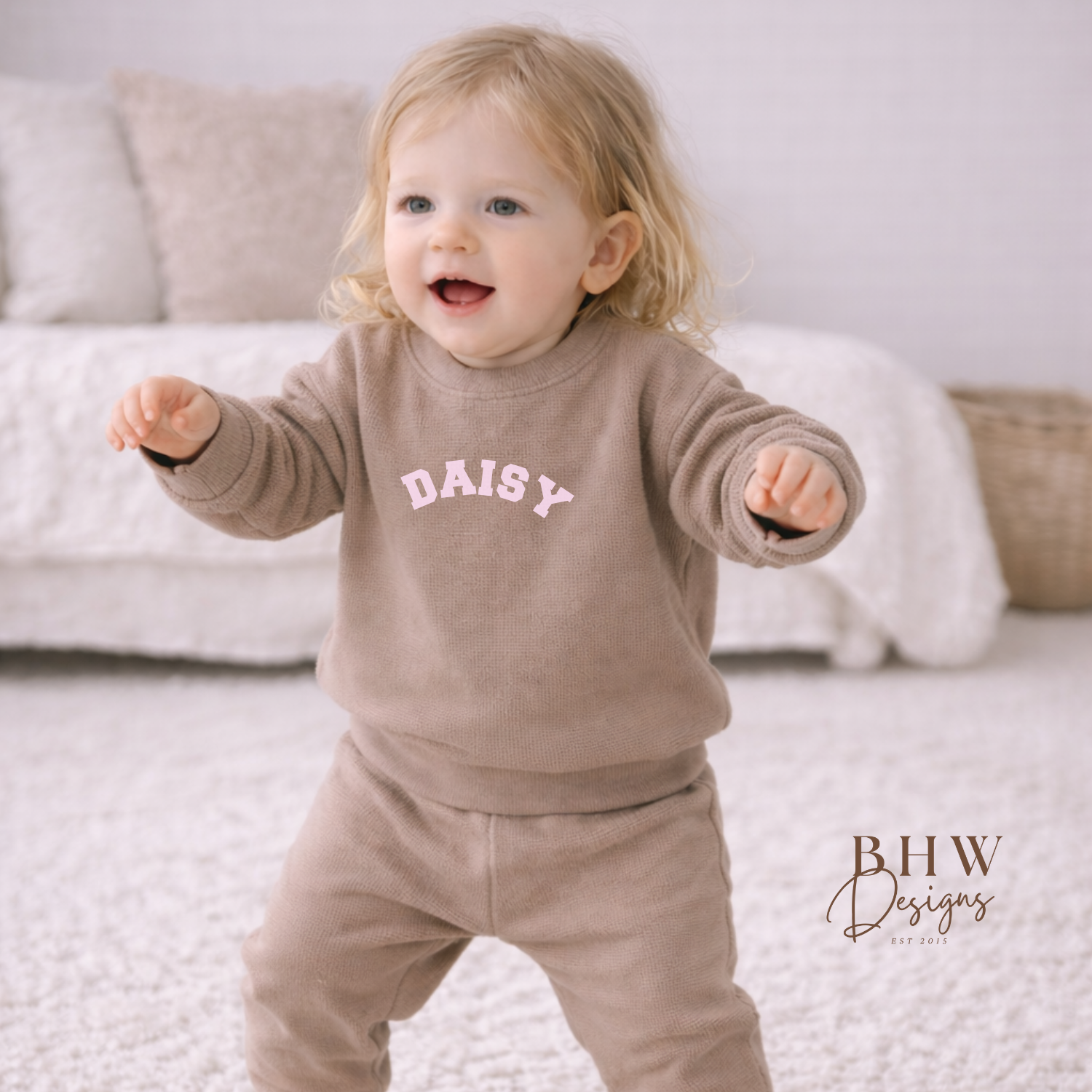 Child wearing a taupe waffle outfit with 'DAISY' printed on it, standing in a room with a couch and basket in the background.