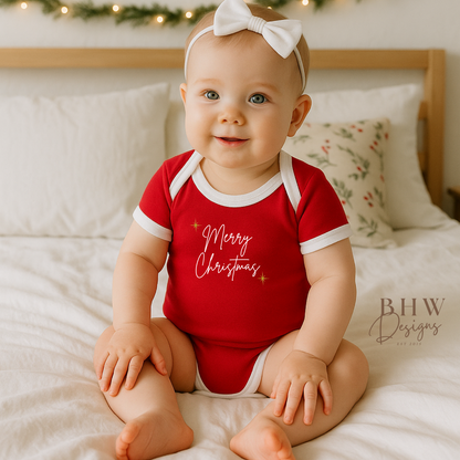 Baby wearing a red 'Merry Christmas' onesie sitting on a bed.