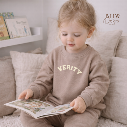 Child wearing a taupe waffle loungewear with 'VERITY' on the front, sitting on a couch reading a book.