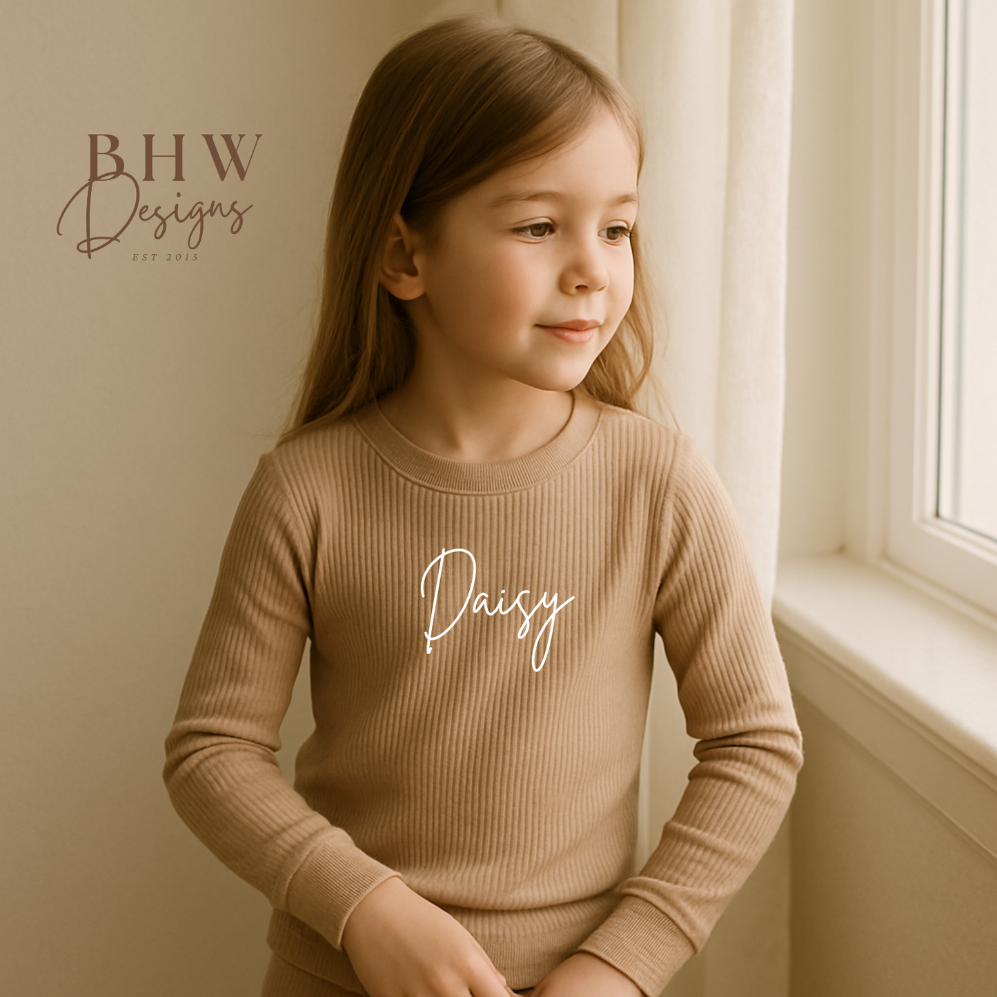 Young girl wearing a beige ribbed sweater with 'Daisy' printed on it, sitting by a window.