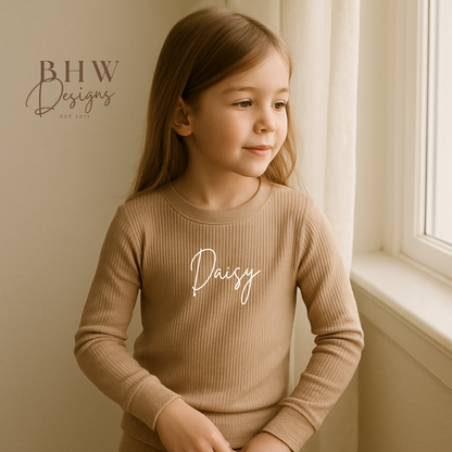 Young girl wearing a beige ribbed sweater with 'Daisy' printed on it, sitting by a window.