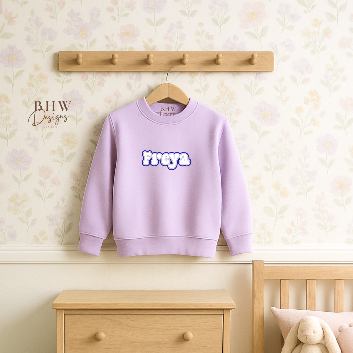 Lavender sweatshirt with 'Freya' text hanging on a wooden coat rack against a light-colored wall.