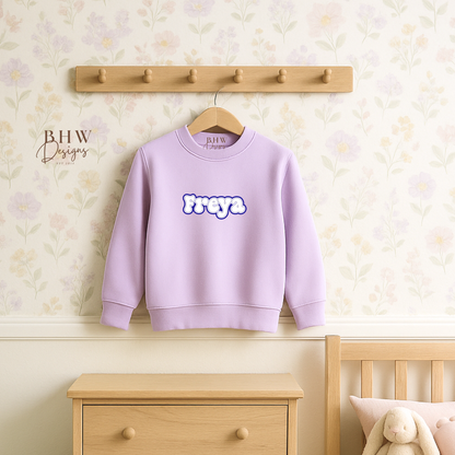 Lavender sweatshirt with 'Freya' text hanging on a wooden coat rack against a light-colored wall.