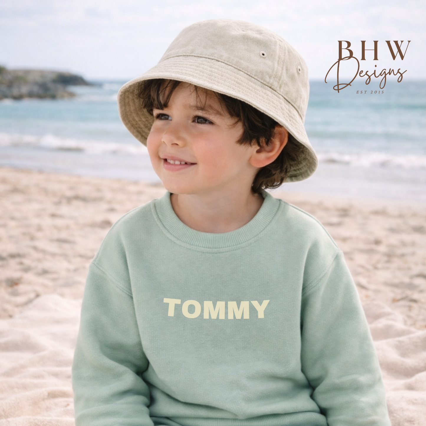 Child wearing a light green 'Tommy' sweatshirt and shorts on a beach, with a sand colour bucket hat