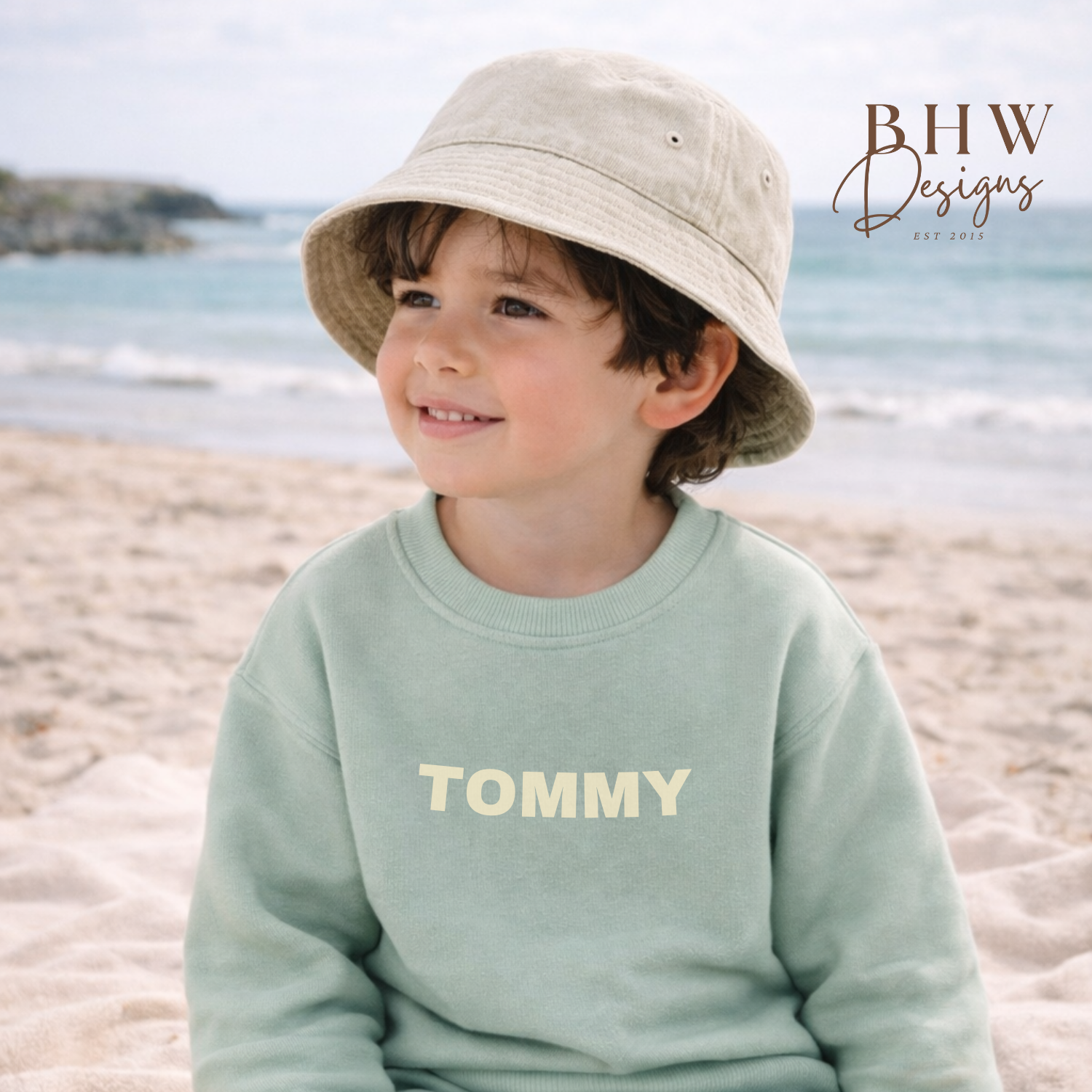 Child wearing a light green 'Tommy' sweatshirt and shorts on a beach, with a sand colour bucket hat