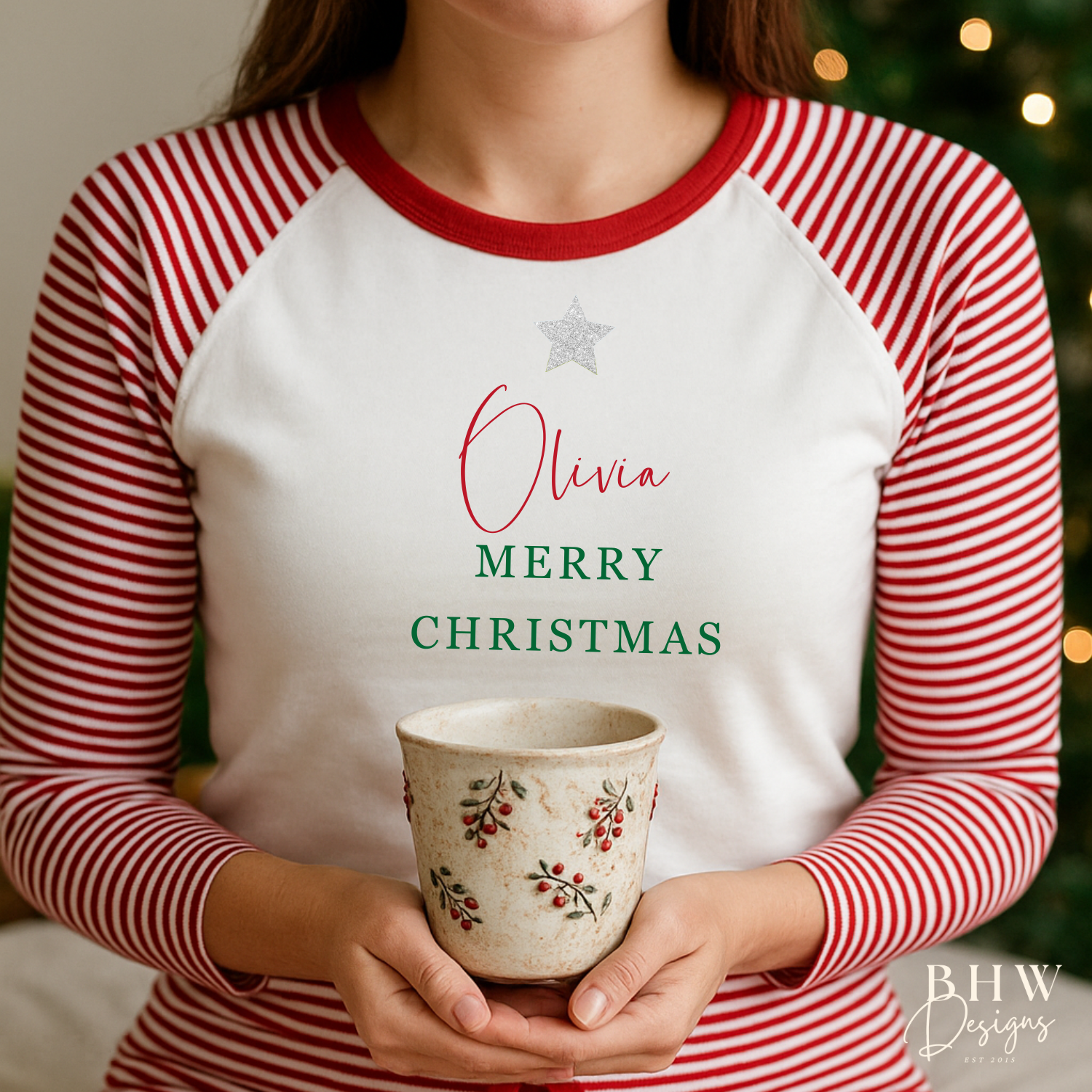Woman wearing red and white striped pyjamas personalised on the front with a silver glitter star and a name in red and merry Christmas in green, sitting on a bed.