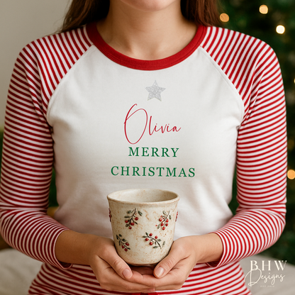 Woman wearing red and white striped pyjamas personalised on the front with a silver glitter star and a name in red and merry Christmas in green, sitting on a bed.