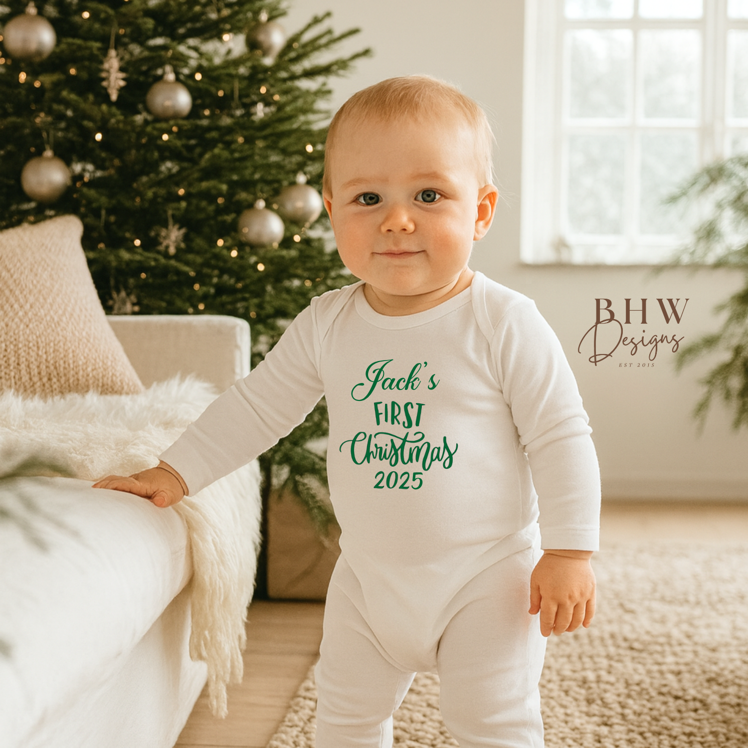 Baby wearing a onesie with 'Jack's First Christmas 2025' text, standing in a room with a decorated Christmas tree.
