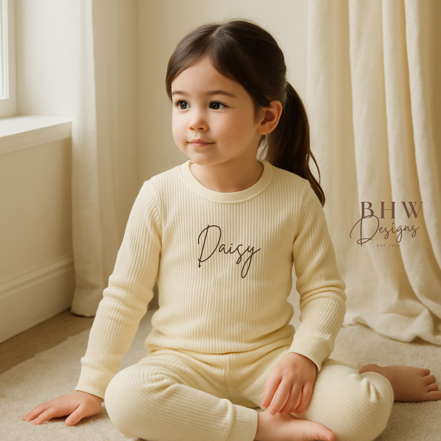 Child wearing a cream-colored outfit with 'Daisy' printed on it, sitting in a softly lit room.
