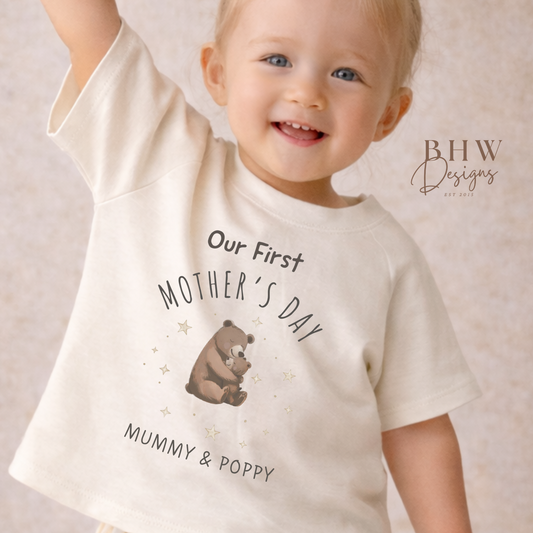 Child wearing a 'Our First Mother's Day' shirt with a bear graphic, standing against a neutral background.