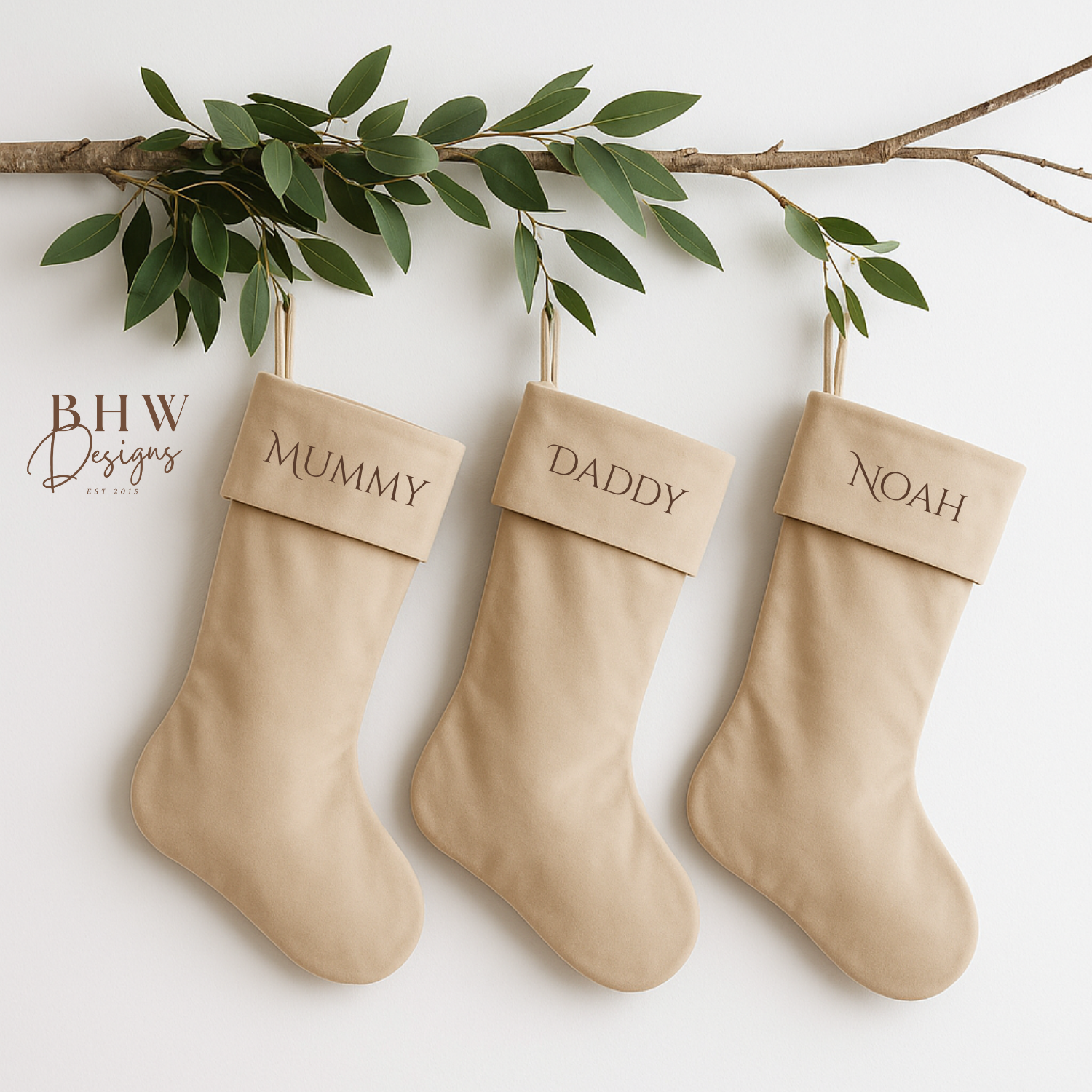 Three beige stockings labeled 'Mummy', 'Daddy', and 'Noah' hanging on a branch with green leaves.