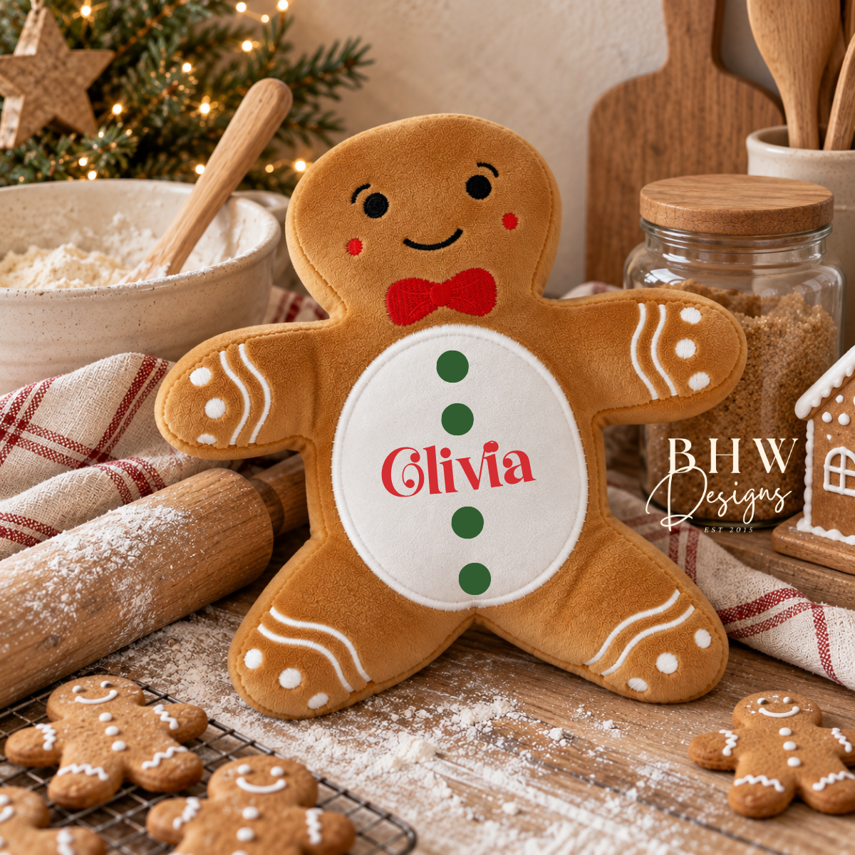A plush gingerbread soft toy, with a white belly panel, red name text reading 'Olivia,' green buttons, a red bow tie, and white icing-style details. The scene is styled on a wooden table with baking props, a small house decoration, and a holiday tree in the background.