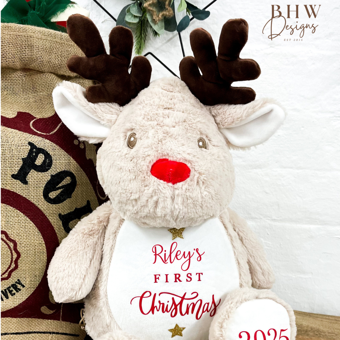 Plush reindeer toy with 'Riley's First Christmas 2025' on the front in red with gold glitter star details. 