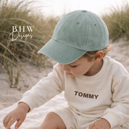 Child wearing a light-coloured shirt with 'Tommy' printed on it and a sage green cap, sitting on sand.