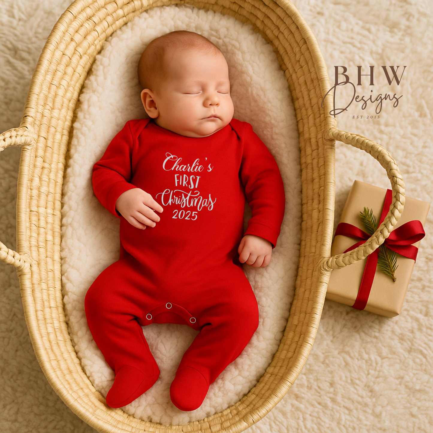 Baby in a red onesie with 'Charlie's First Christmas 2025' text, lying in a woven basket with a gift box.