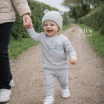 Child in a light grey outfit with 'Noah' printed on it, holding hands with an adult outdoors.