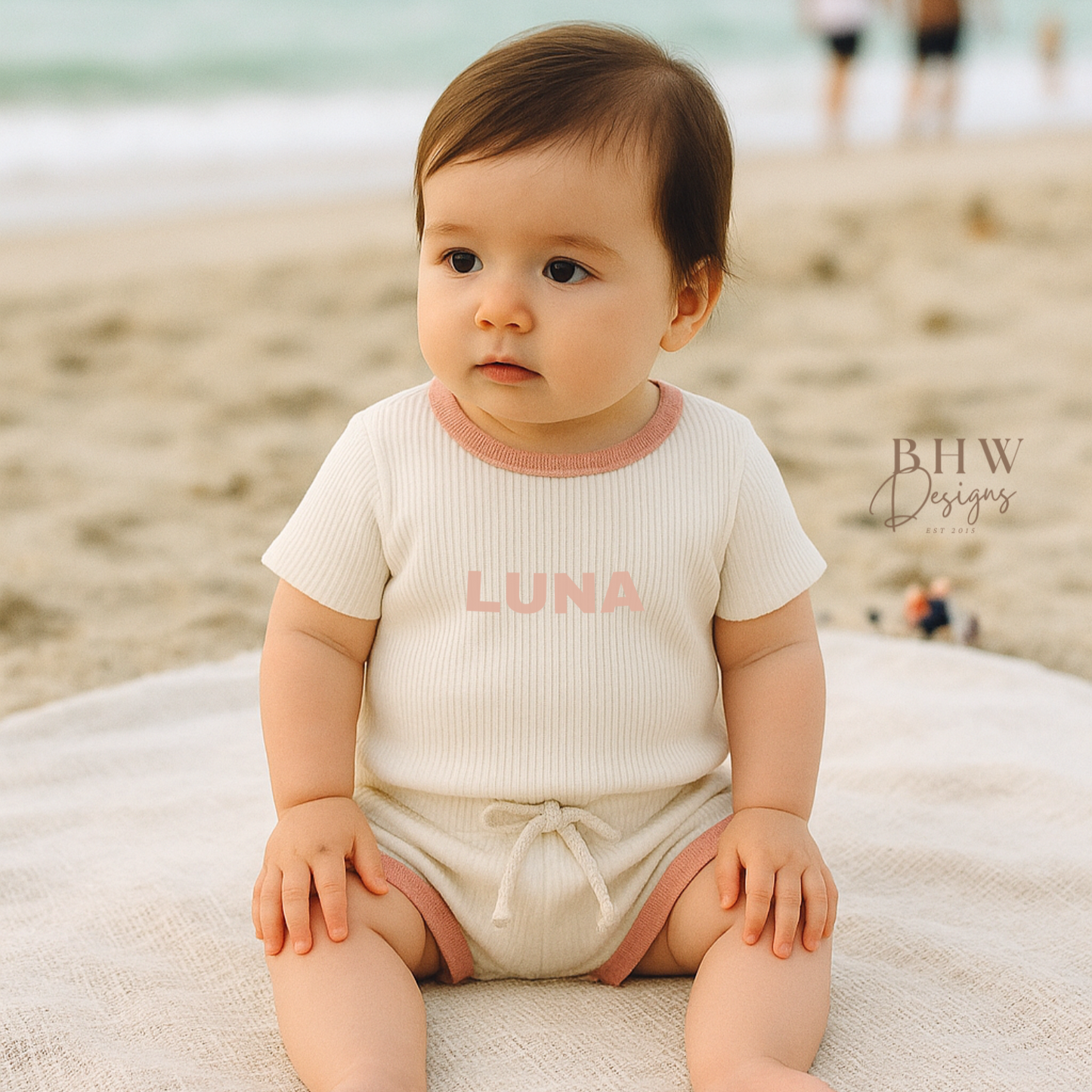 Baby sitting on a towel at the beach wearing a white ribbed outfit with 'LUNA' on the front