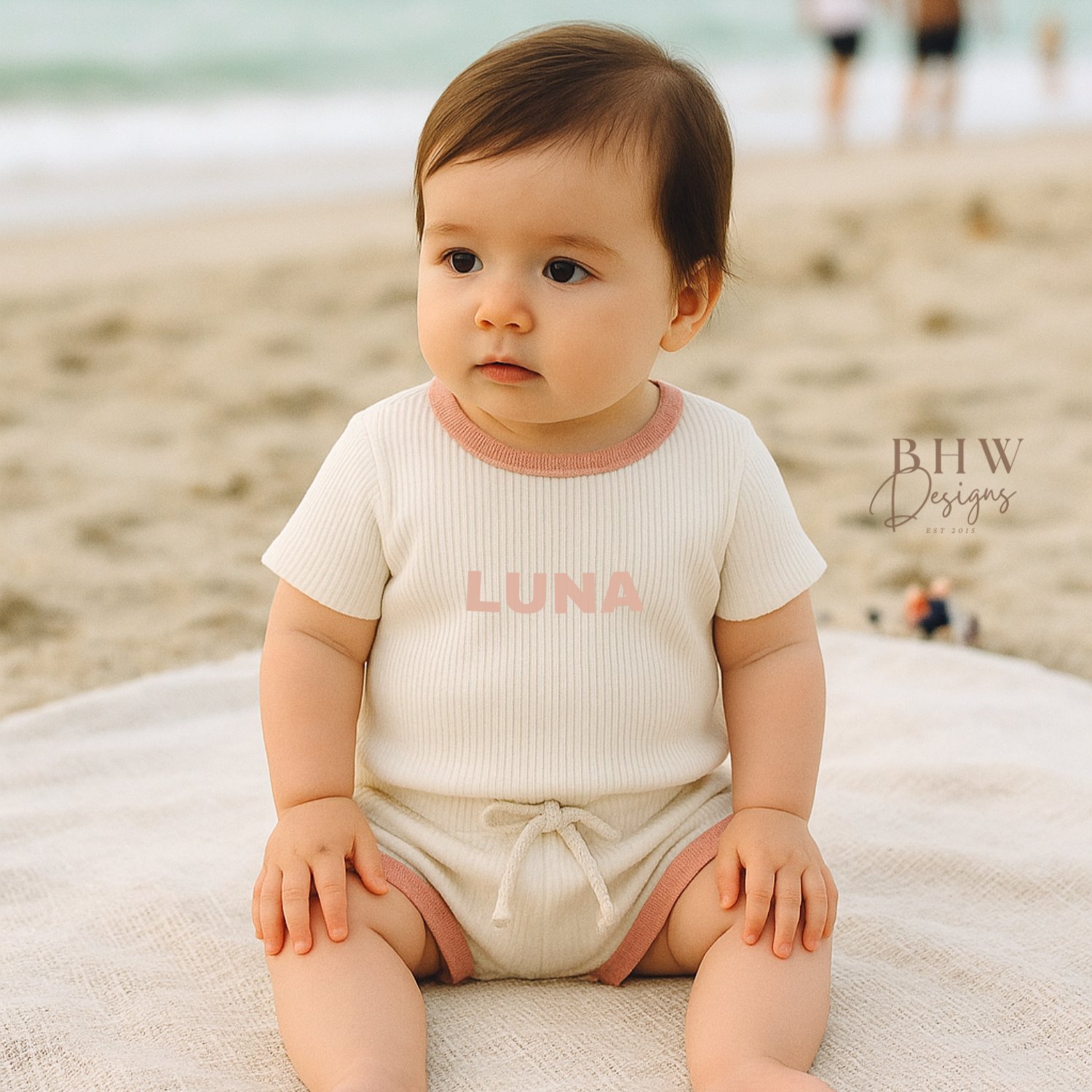 Baby sitting on a towel at the beach wearing a white ribbed outfit with 'LUNA' on the front
