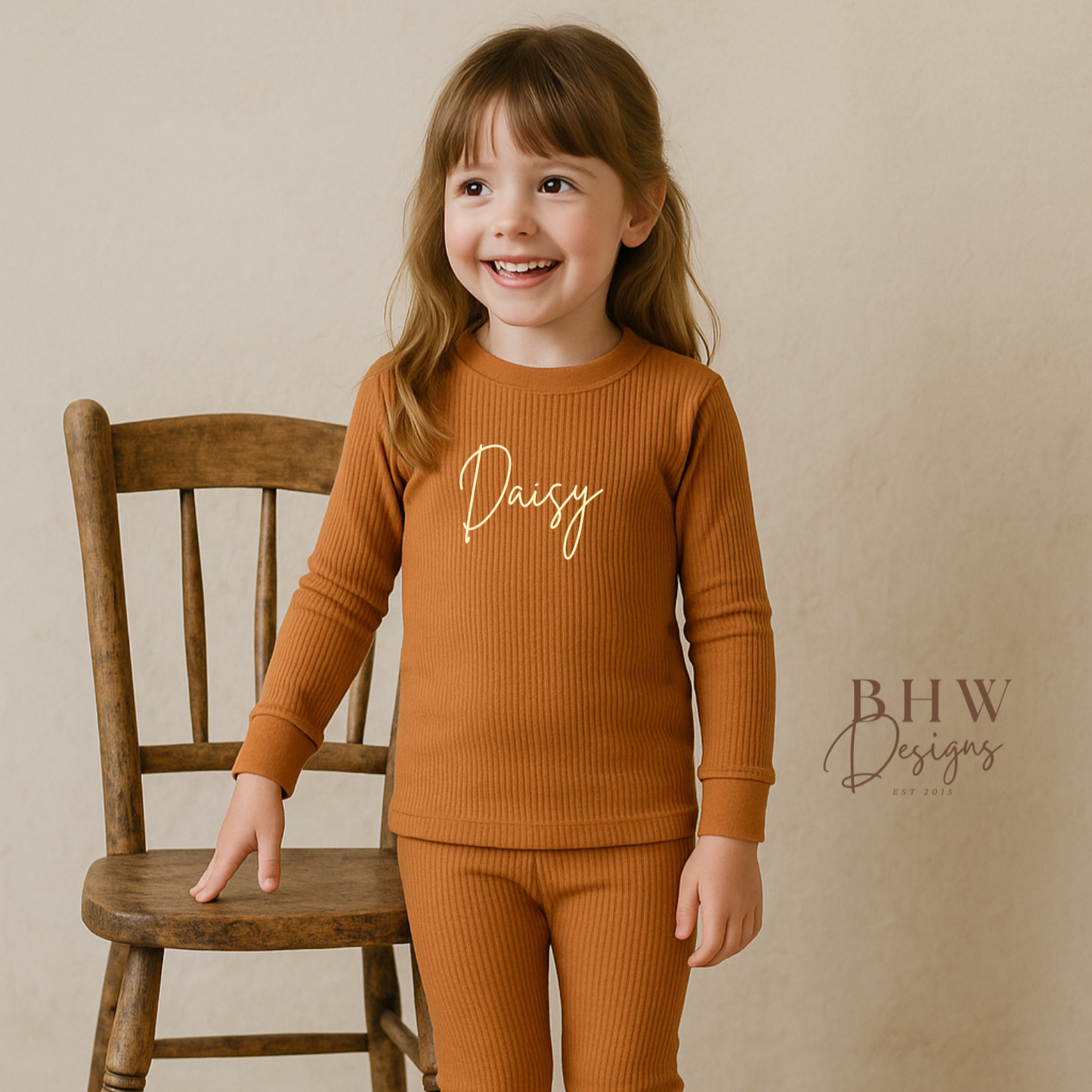 Girl wearing a rust outfit with 'Daisy' printed on it, standing next to a wooden chair against a beige background.
