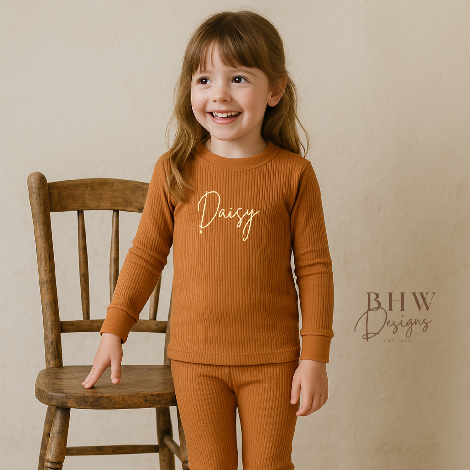 Girl wearing a rust outfit with 'Daisy' printed on it, standing next to a wooden chair against a beige background.