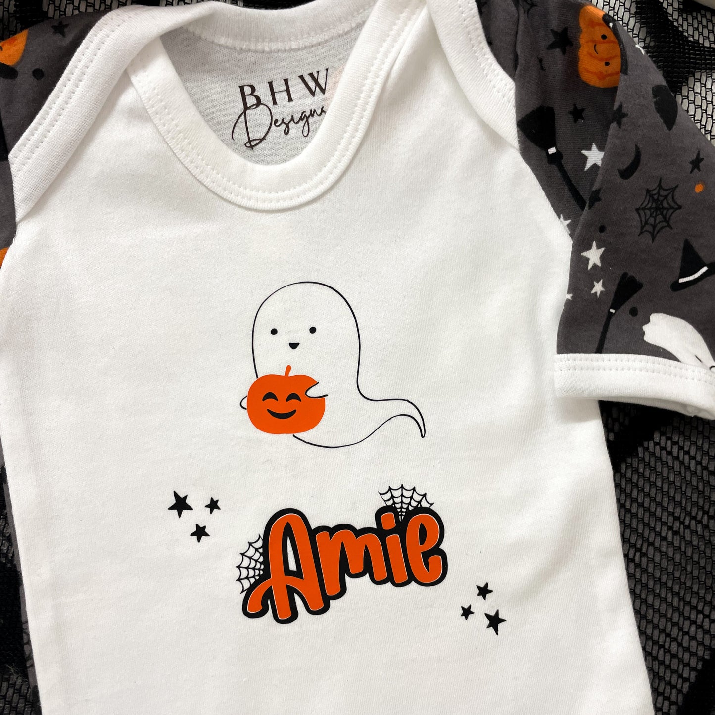 White baby onesie with ghost and pumpkin design, featuring the name 'Amie', on a black and gray patterned background.