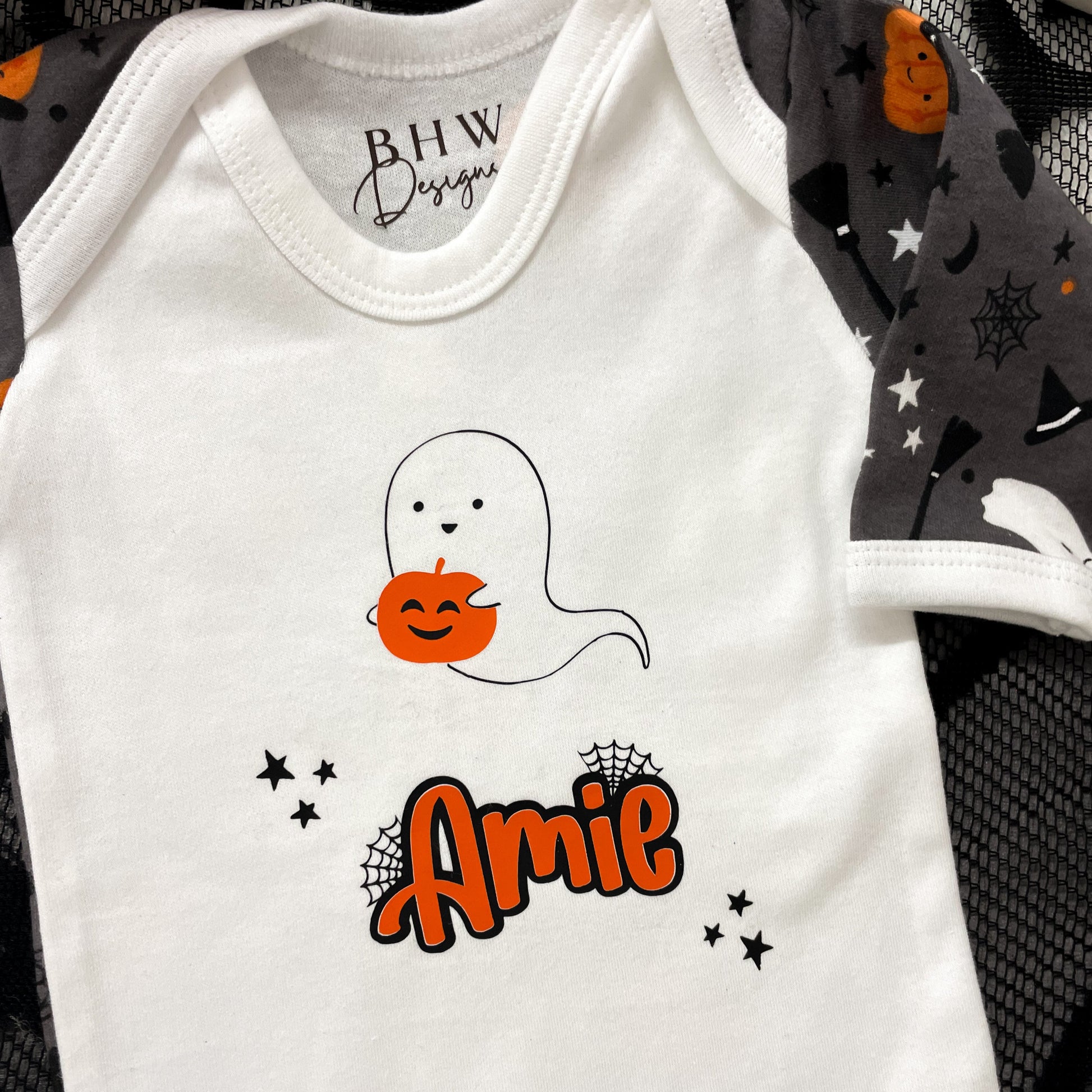 White baby onesie with ghost and pumpkin design, featuring the name 'Amie', on a black and gray patterned background.