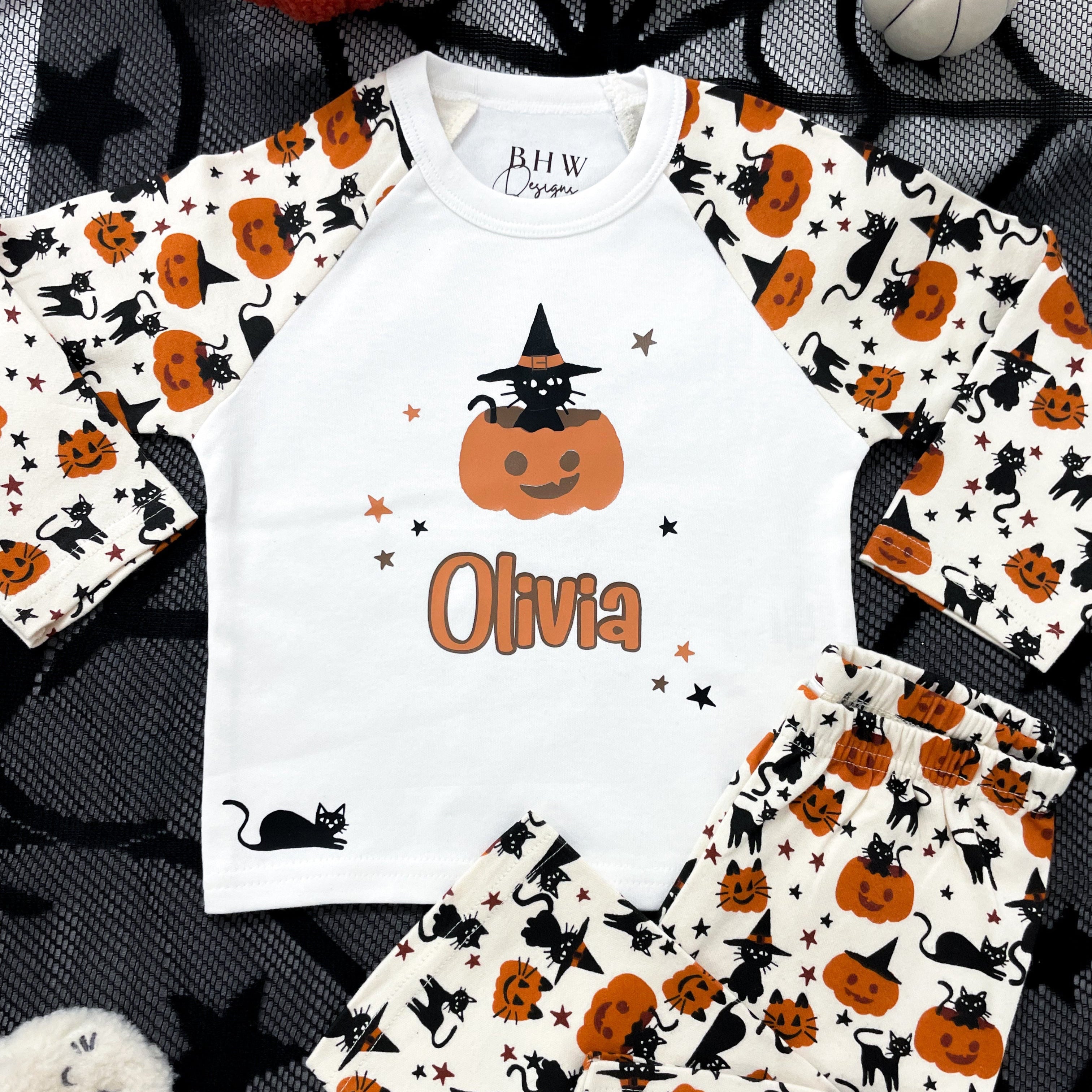 Children's Halloween-themed pyjamas with a black cat and pumpkin pattern, personalised name 'Olivia' printed on the front, displayed on a dark background with Halloween decorations.