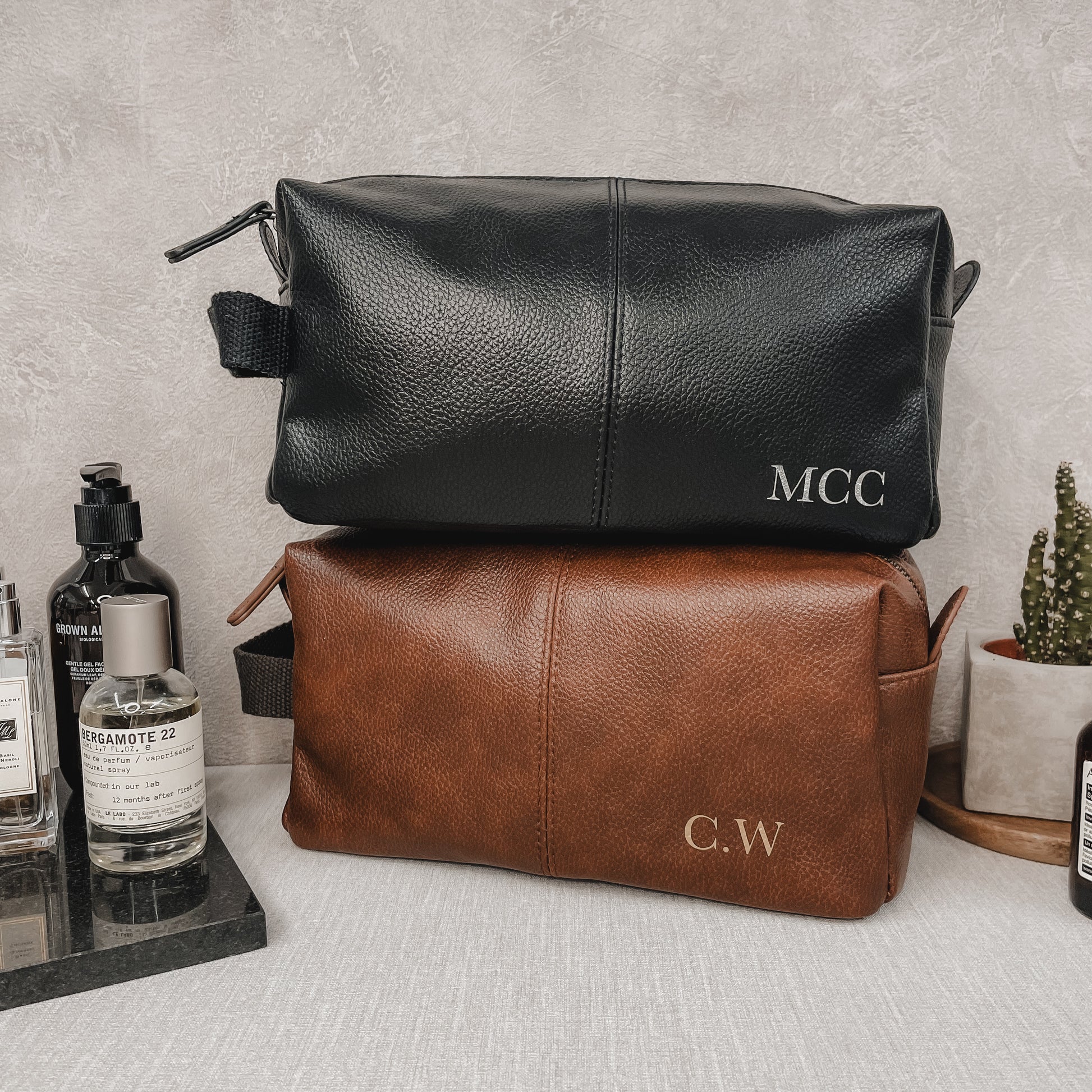 Black and brown faux leather toiletry bags with initials on a grey surface.