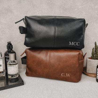 Black and brown faux leather toiletry bags with initials on a grey surface.